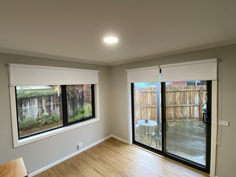 White blockout roller blinds fitted to a window and sliding glass door in a bright room, with a view to a courtyard garden