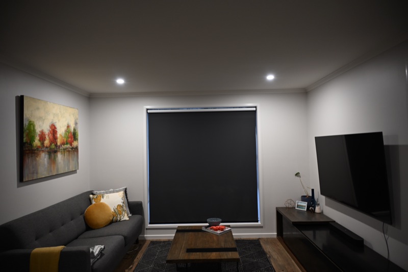 Charcoal blockout roller blind fitted into a window reveal in a modern living room, with sofa and wall-mounted television visible