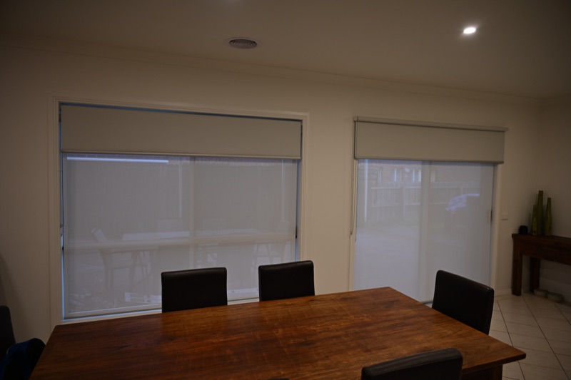 White dual roller blinds installed on a side window and sliding glass door in a dining room, looking out to a garden