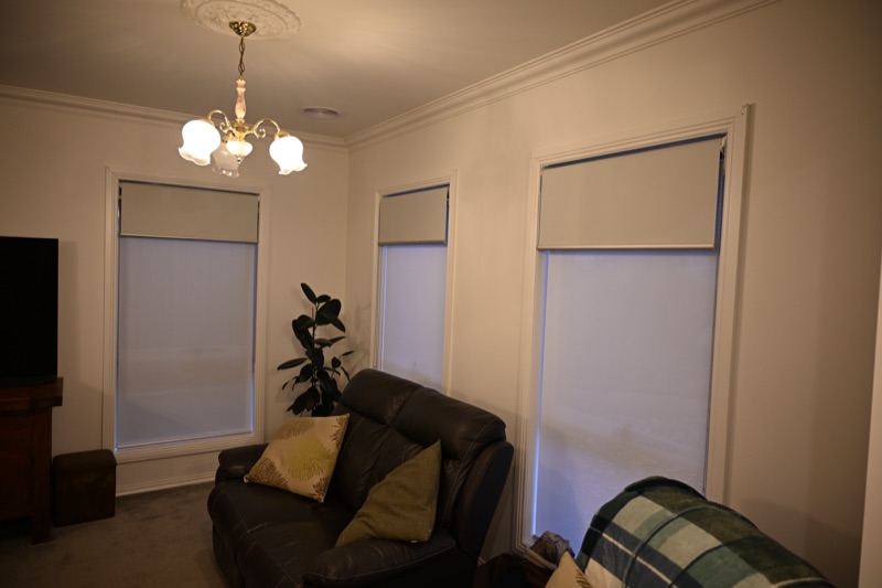 Three cream dual roller blinds fitted in ornate white-framed windows of a traditional lounge room with leather sofa and chandelier